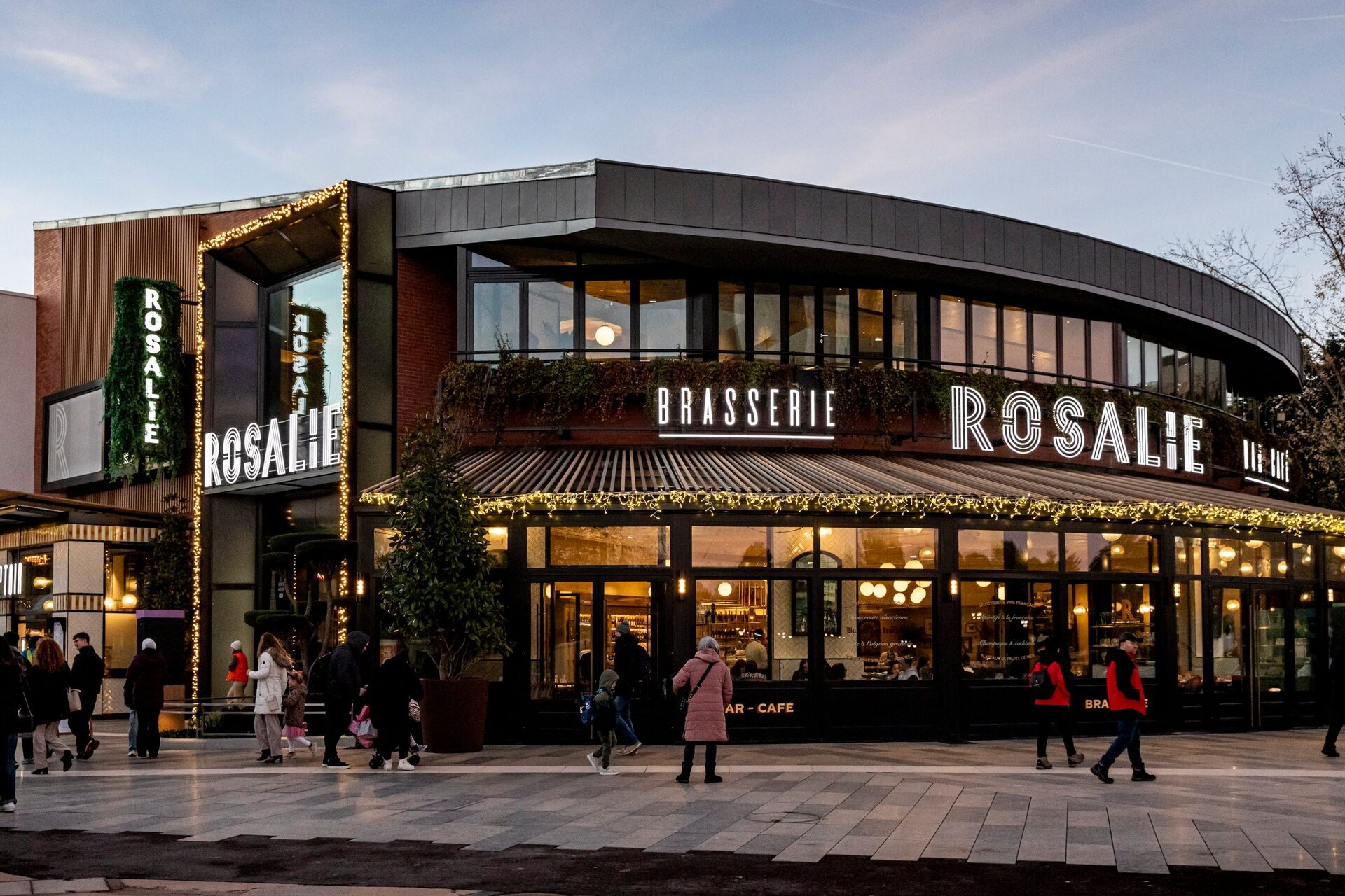Façade illuminée de la Brasserie Rosalie à Disney Village au crépuscule, avec des passants devant sa verrière. Photo : © Rosalie / Simon Detraz. Façade illuminée de la Brasserie Rosalie à Disney Village au crépuscule, avec des passants devant sa verrière. Photo : © Rosalie / Simon Detraz.