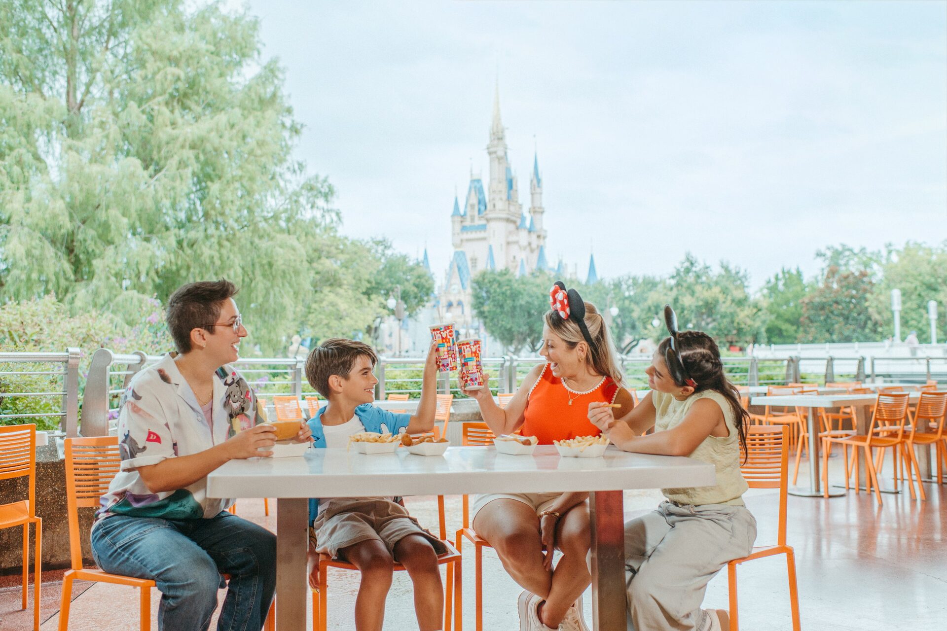 Four guests raise their drinks around an orange terrace table, Cinderella Castle rising beyond the trees, at Magic Kingdom Four guests raise their drinks around an orange terrace table, Cinderella Castle rising beyond the trees, at Magic Kingdom