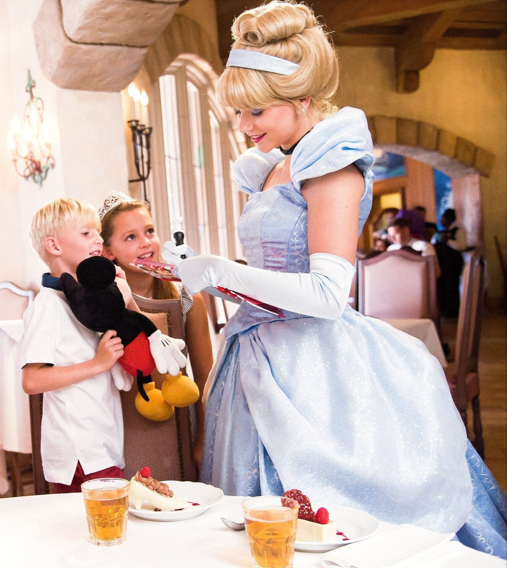 Cinderella greets two children at their table at Auberge de Cendrillon in Disneyland Park, one of them hugging a Mickey plush. Photo: ©Disney.