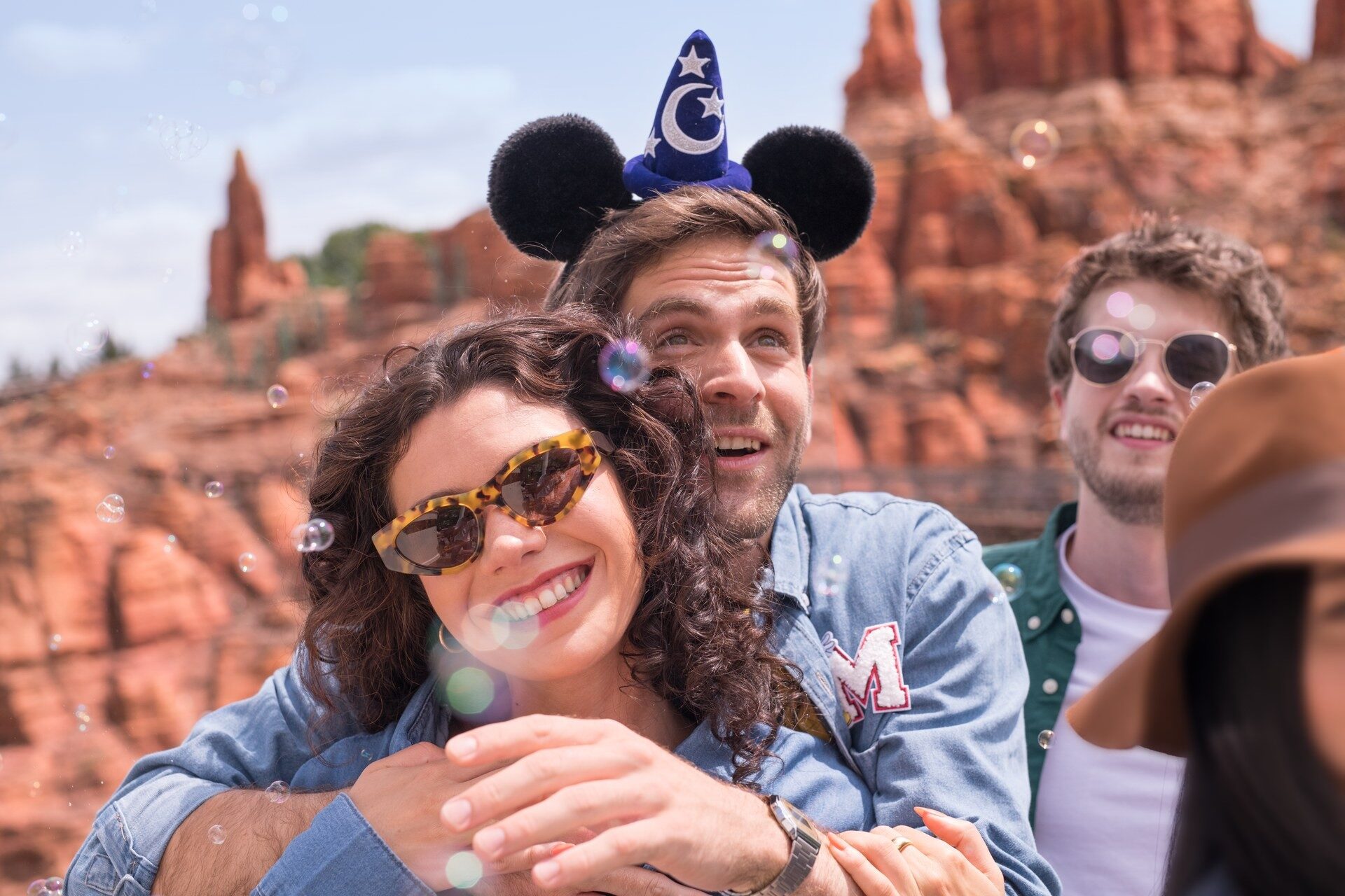 Deux visiteurs souriants, dont un coiffé d'un chapeau à oreilles de Mickey, devant les rochers rouges de Big Thunder Mountain. Photo : ©Disney. Deux visiteurs souriants, dont un coiffé d'un chapeau à oreilles de Mickey, devant les rochers rouges de Big Thunder Mountain. Photo : ©Disney.