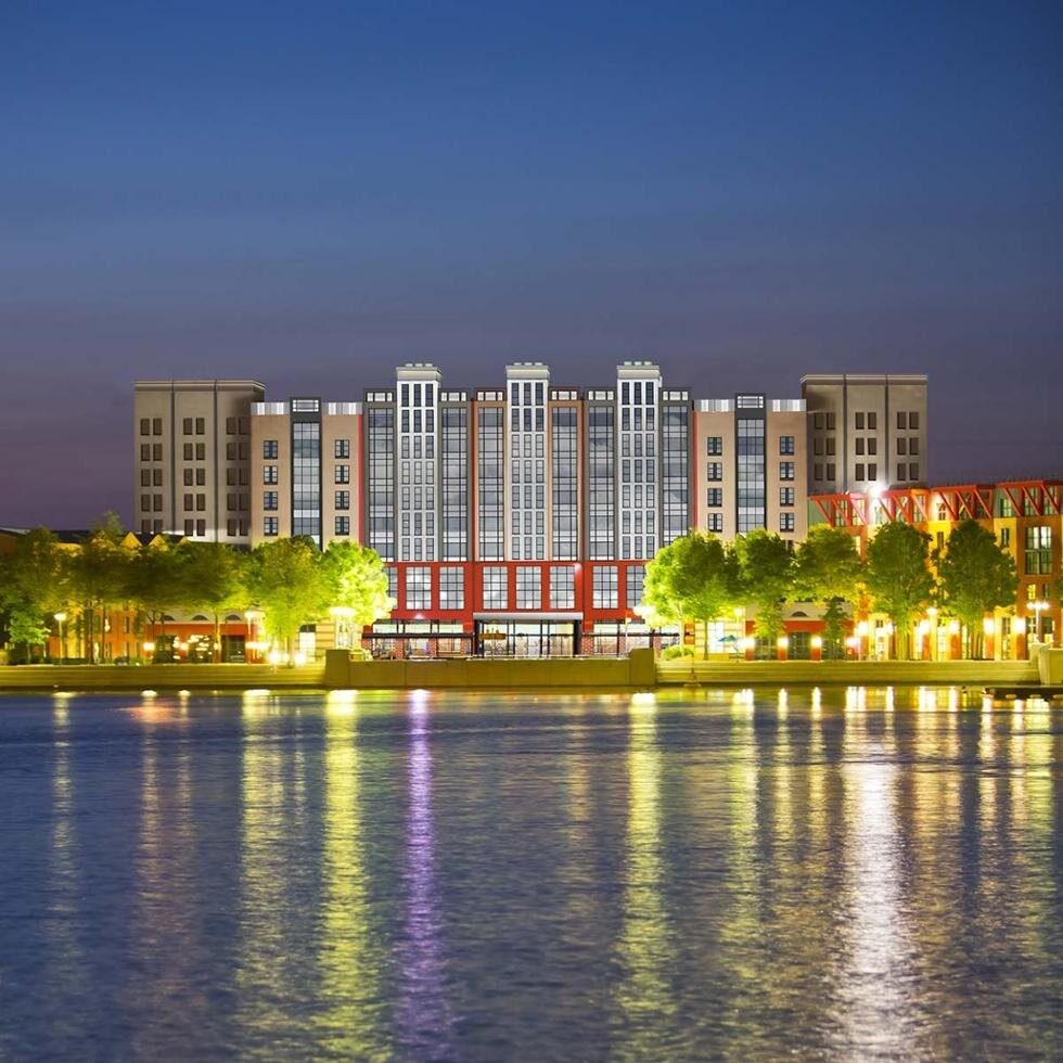 La façade du Disney Hotel New York – The Art of Marvel à la tombée du jour, avec ses gratte-ciel illuminés qui se reflètent dans le lac Disney. Photo : ©Disney. La façade du Disney Hotel New York – The Art of Marvel à la tombée du jour, avec ses gratte-ciel illuminés qui se reflètent dans le lac Disney. Photo : ©Disney.