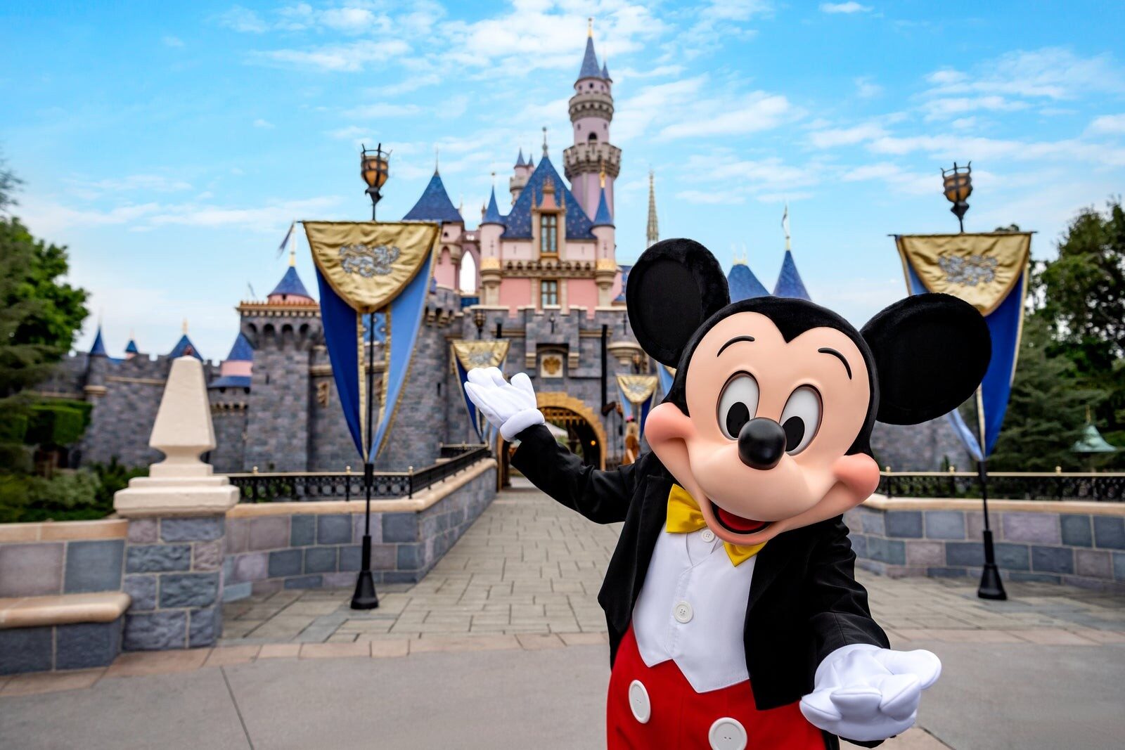 Mickey Mouse waving in front of Sleeping Beauty Castle and its blue banners Mickey Mouse waving in front of Sleeping Beauty Castle and its blue banners